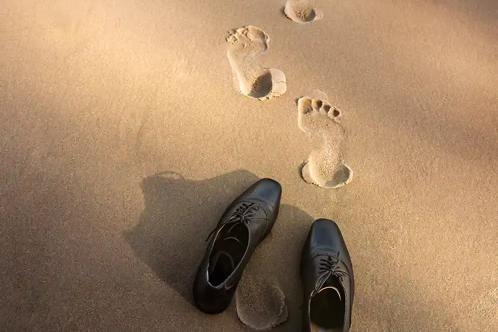 Business shoes left on beach with bare footprints walking away from them while on a bleisure trip.